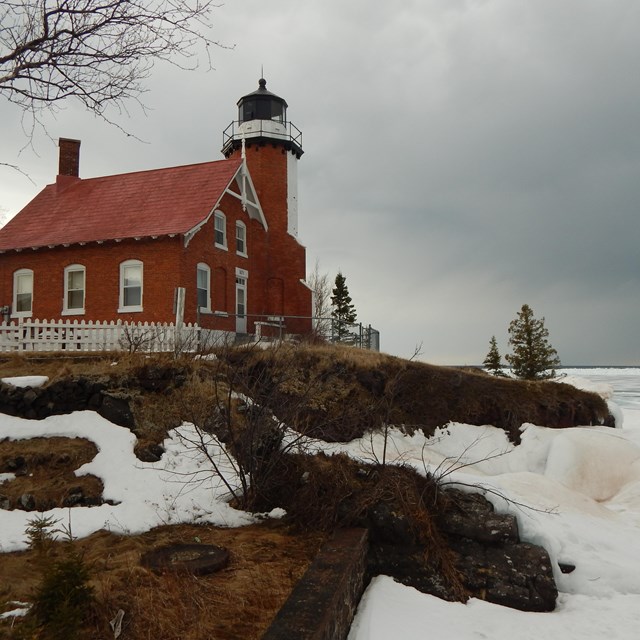 A brick lighthouse stands tall on a lakeshore over a rocky outcropping covered in snow and ice.