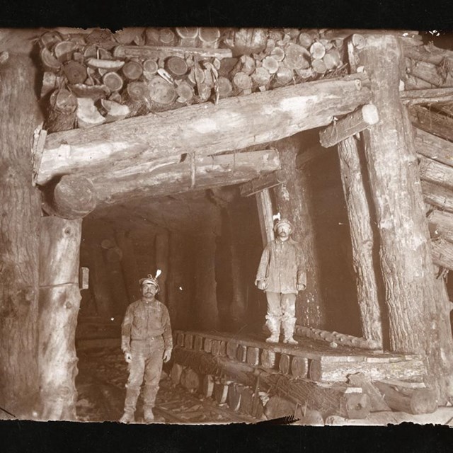In this historic black and white image, two men stand under massive wood supports underground.