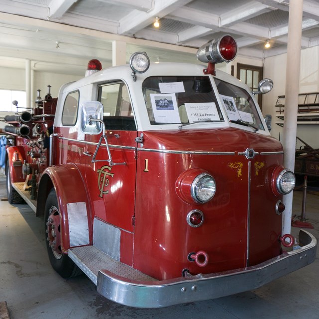 An older red fire truck parked in a historic firehouse. White posts support the upper floor. 