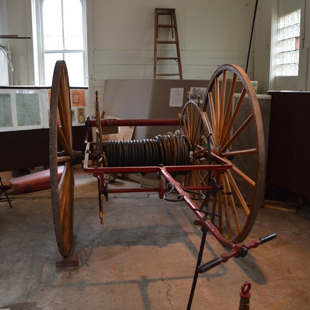 A historic fire hose reel with large wood wheels sits on a hardwood floor.