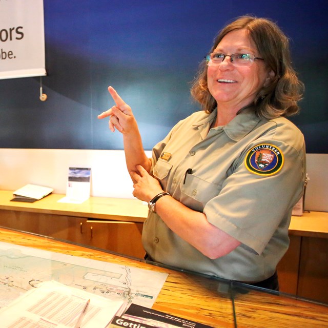 A person wearing an NPS volunteer uniform signs with a visitor at an information desk.