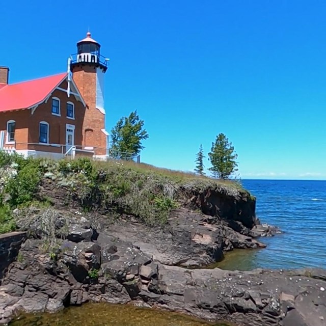 A red brick lighthouse sits atop a rocky shoreline. A lake etends off to the right under blue skies.
