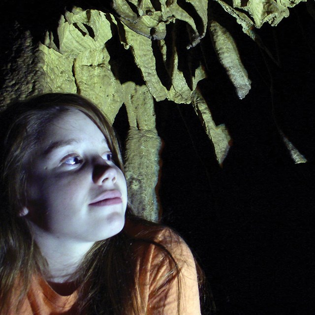 Girl in orange shirt looks up and shadows on the wall of a cave.