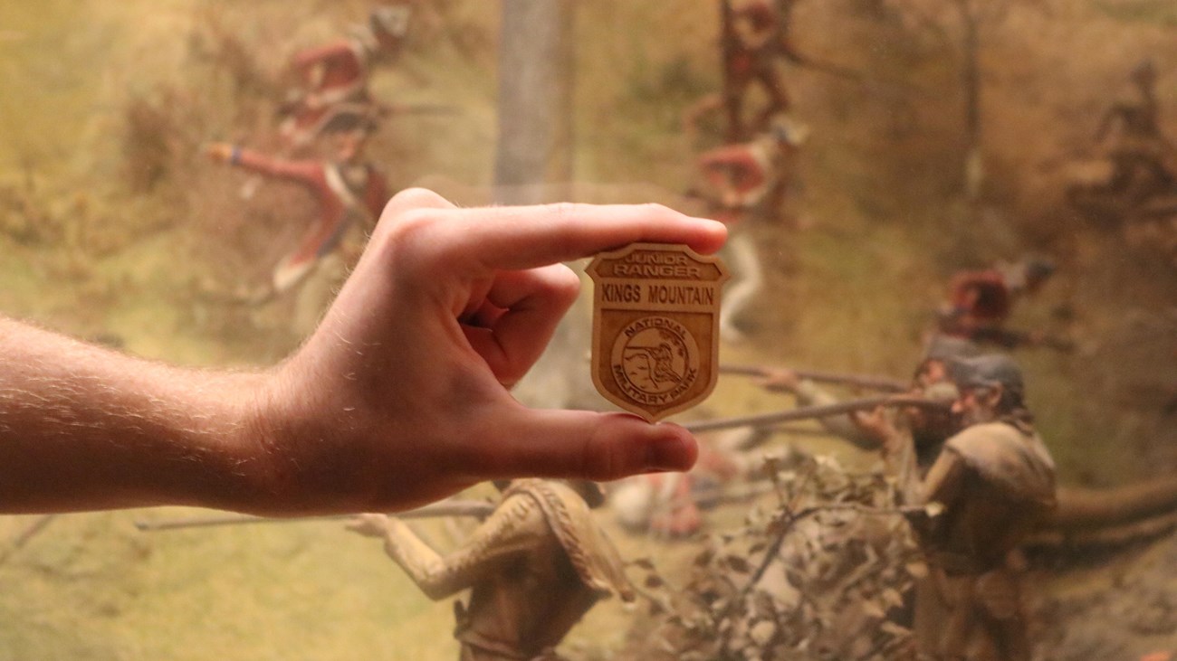 A hand holding a wooden badge for Kings Mountain and an etching of a soldier firing a musket.