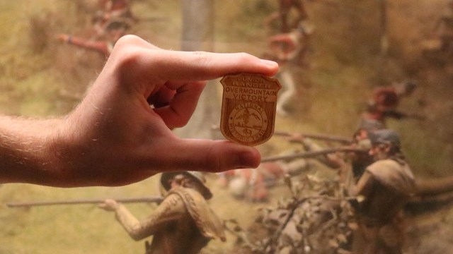 A hand holds a wooden badge that has Overmountain Victory National Historic Trail on it