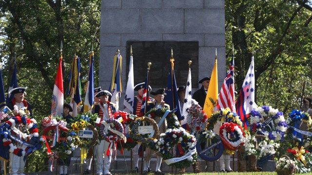 Living historians line up by wreaths with flowers by a large stone monument.