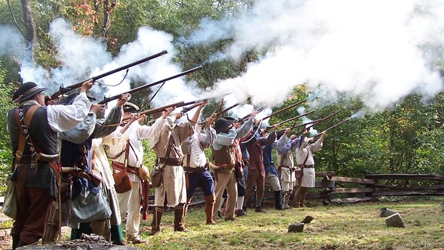 An image of a park sign and a historical marker along a road with trees in the background.