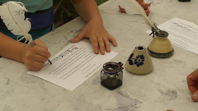 Living historians fire muskets while laying on rocks with trees in the background.