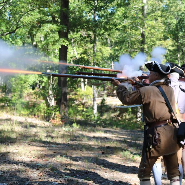 Image shows men wearing Rev War clothing standing in a line firing muskets.