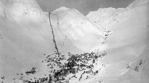 Black and white photo of people gathered in a flat area before a steep line ascends a snowy pass.