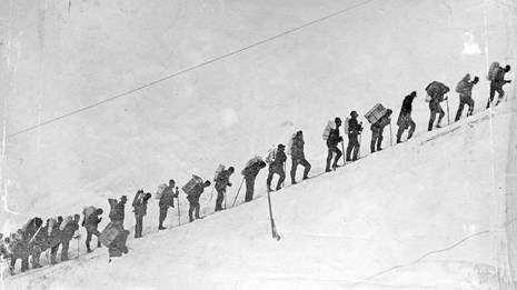 Black and white photo of people standing in a line on a snowy slope.
