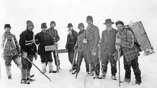 Black and white photo of 9 bundled men standing at the Alaska-British Columbia border in the snow.