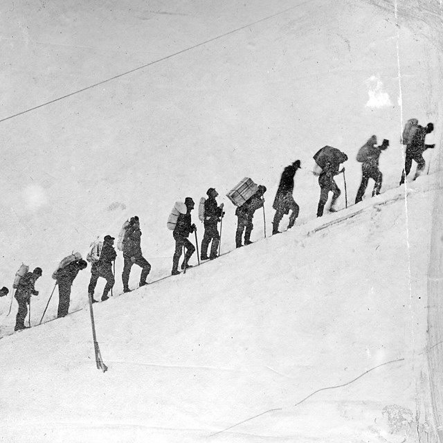Black and white photo of people standing in a line on a snowy slope.
