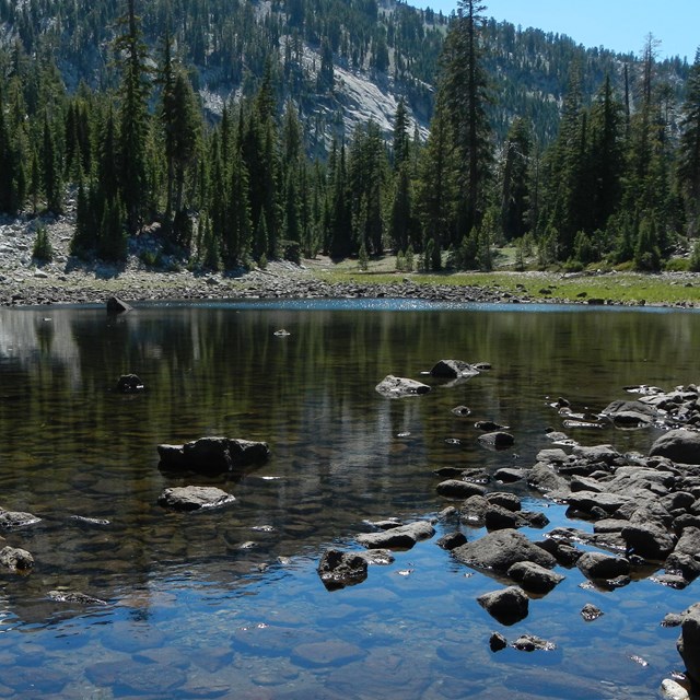 Shallow lake in Lassen Volcanic NP with rocks in foreground and trees and cliff along the shore