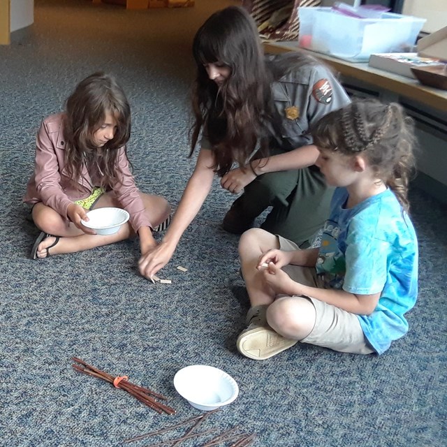 4 kids and female ranger sit in circle on floor playing a game with bowls, sticks, and wooden chips.