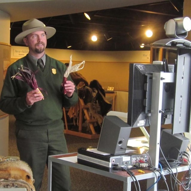 Male ranger holds 2 objects while looking at a tv monitor.