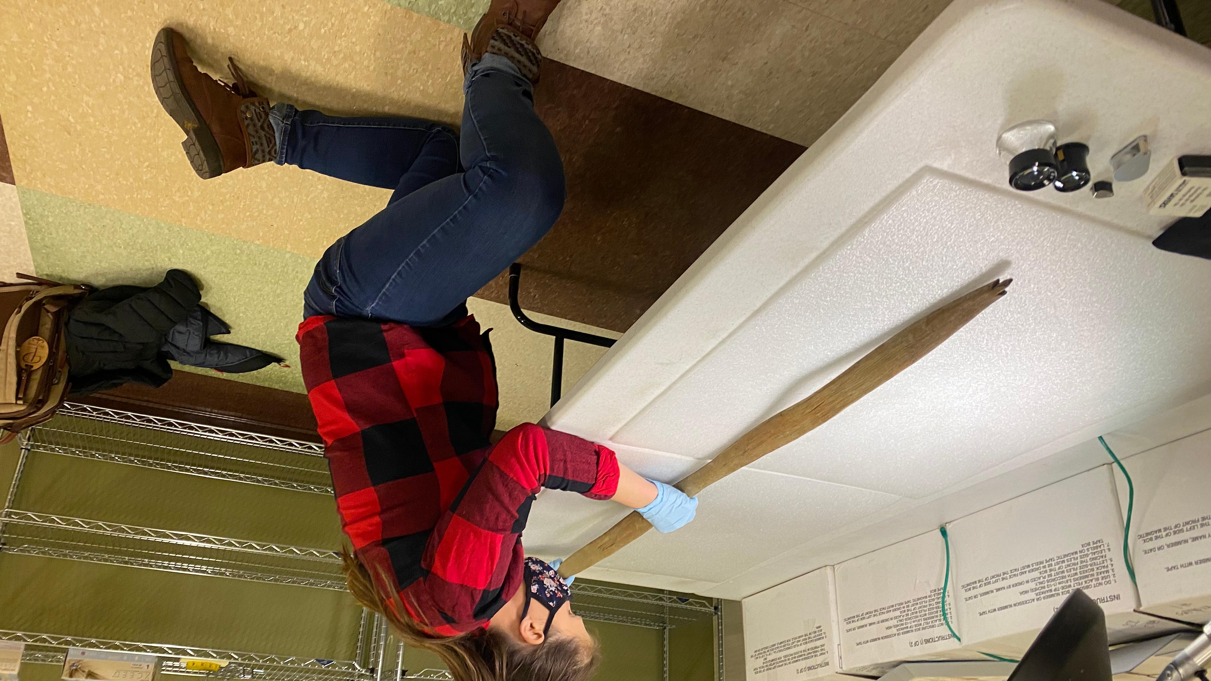 a women in a red flannel inspects a handcrafted wooden hunting bow on a table