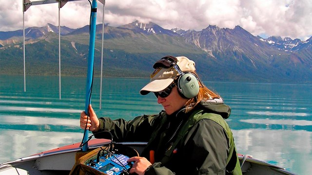 A woman holding a large antenna sits in a boat on a lake surrounded by colorful mountains.