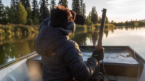 A hunter reaches holds a rifle while travelling in a boat