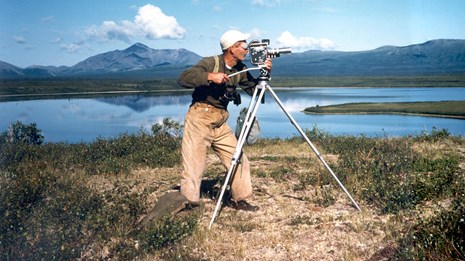 Vintage photo of a man filming with an old movie camera on a tripod near a blue lake and mountains.