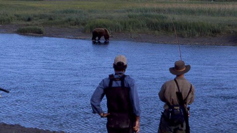 Two fishermen watch a brown bear on the opposite side of the river.