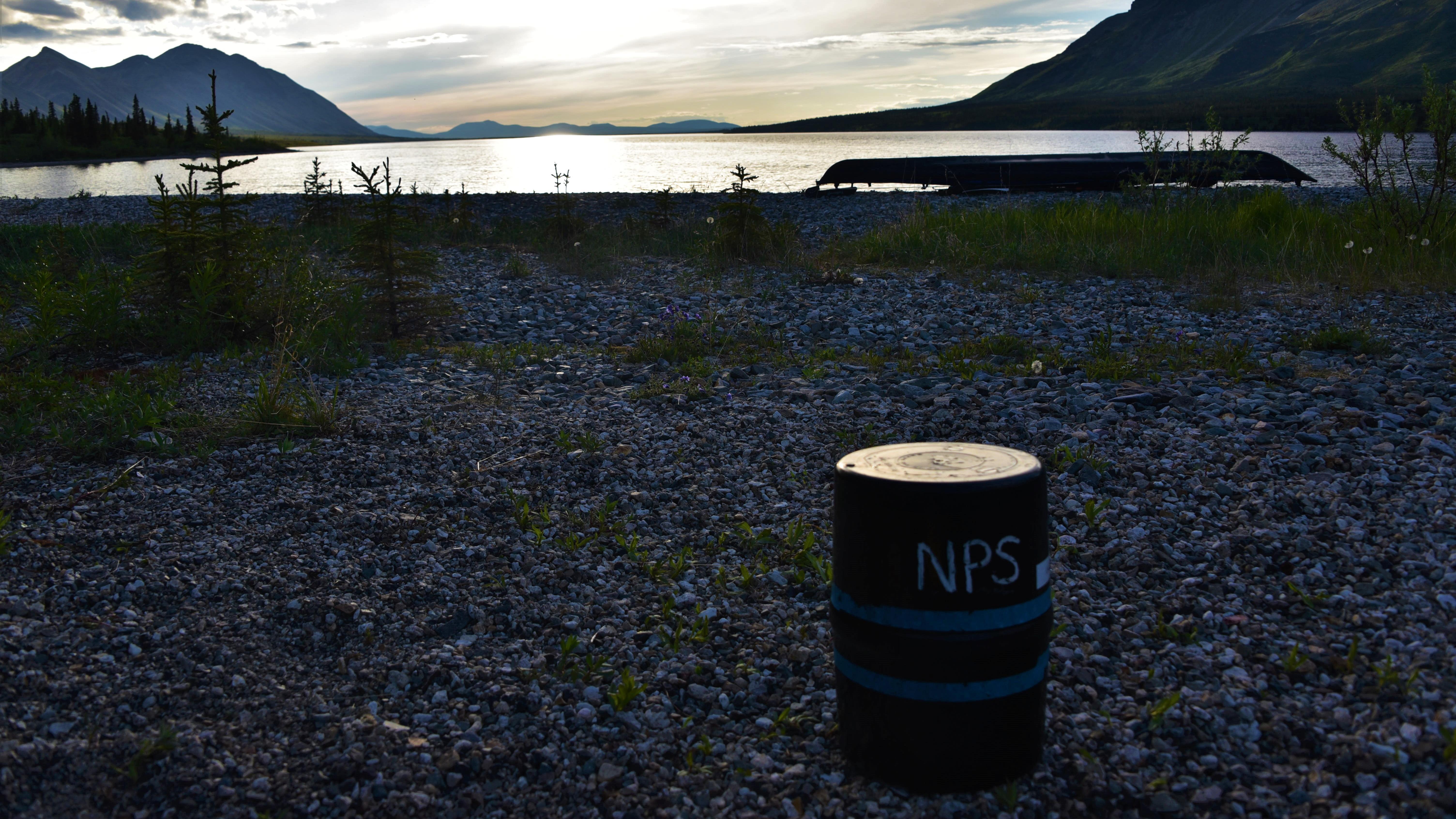 a bear barrel sits on a beach with distant kayak and mountains at dusk in the background