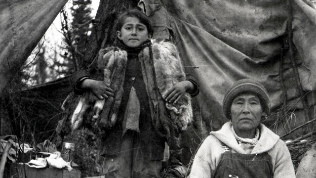 Historic photo of woman and child in front of tent with fish and furs in foreground.