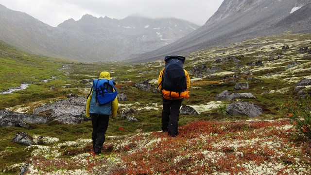 Two people with backpacks hike through mountainous tundra
