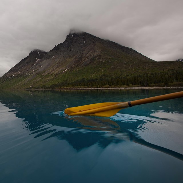 photo of a kayak paddle breaking the water's surface in a blue lake with a mountain in the distance.