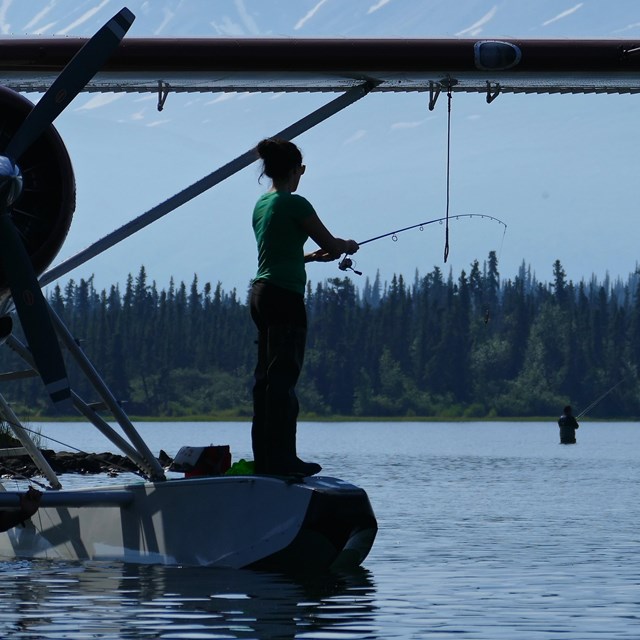 An angler fishes while standing on the float of a plane on a lake with mountains in the background