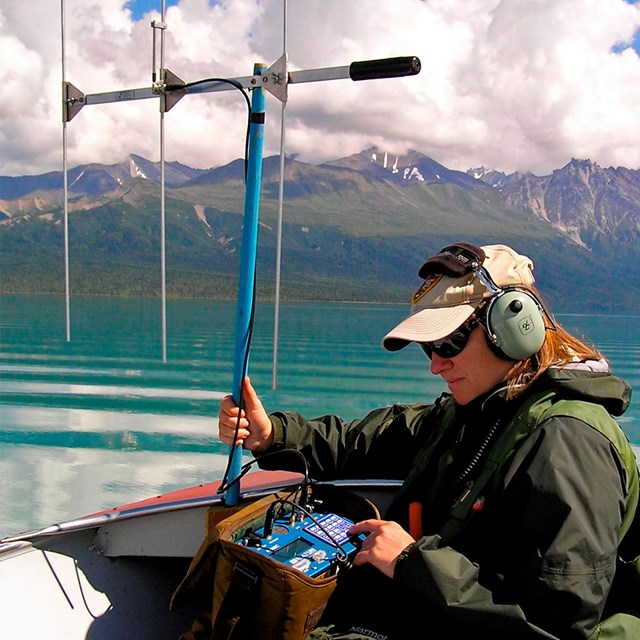 A woman holding a large antenna sits in a boat on a lake surrounded by colorful mountains.