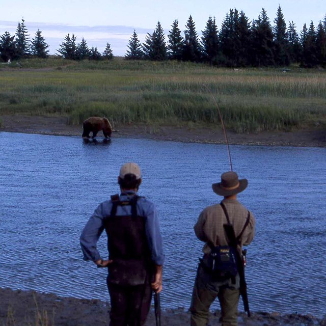 Two fishermen watch a brown bear on the opposite side of the river.