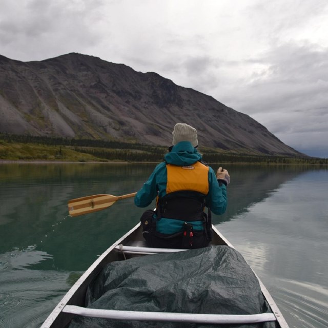 A women with a yellow PFD paddles a canoe on a calm, reflecting lake