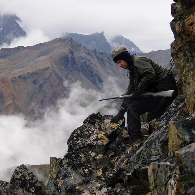 A man with a rifle sits on a steep, rocky slope