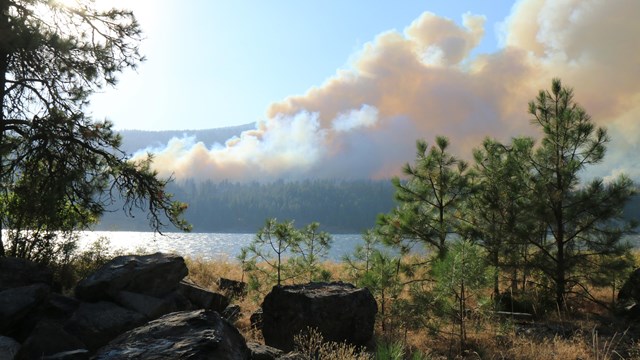 Smoke billowing on a hillside overlooking a lake and trees.
