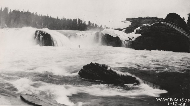 Black and white image of a rushing waterfall with large rocks in the river.