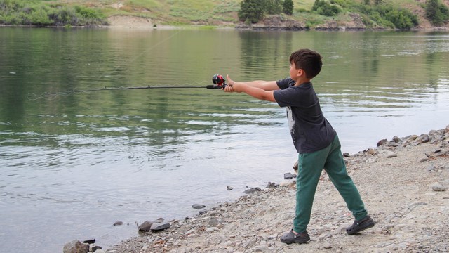 A young boy with a black shirt casts a fishing pole out towards the lake water.