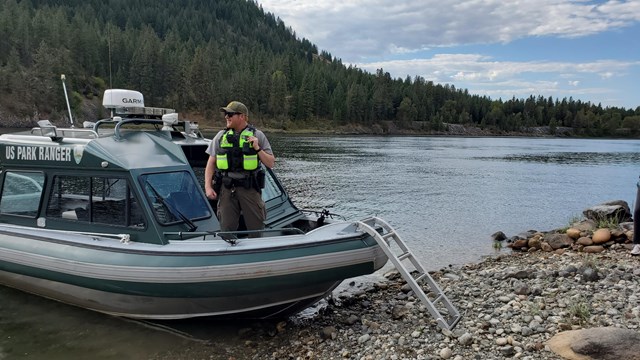 Park ranger in a life vest stands on a boat that is docked on the shoreline of the lake.
