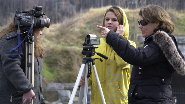 Two video cameras are set up and a woman in a black jacket points into the distance.