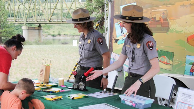 Two park rangers in grey shirts direct visitors to an activity on a table with a green table cloth.