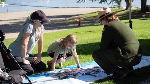 A ranger squats down children and parent and helps them with a fish puzzle.