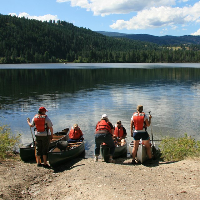 Group of adults with red life vests are launching canoes into a shiny blue lake.