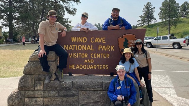 six people smiling and posing with a sign that reads 'wind cave national park visitor center and hea