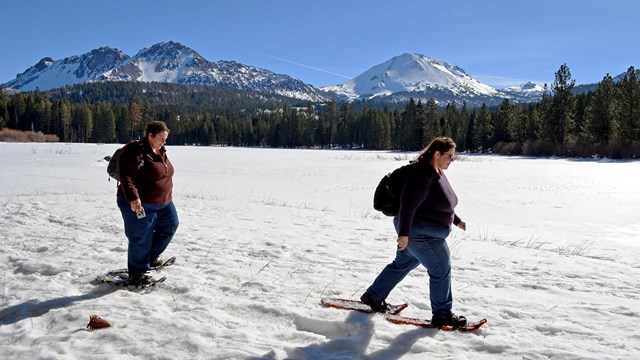 Two women on snowshoes walk across the shore of a snow-covered lake backed by volcanic peaks.