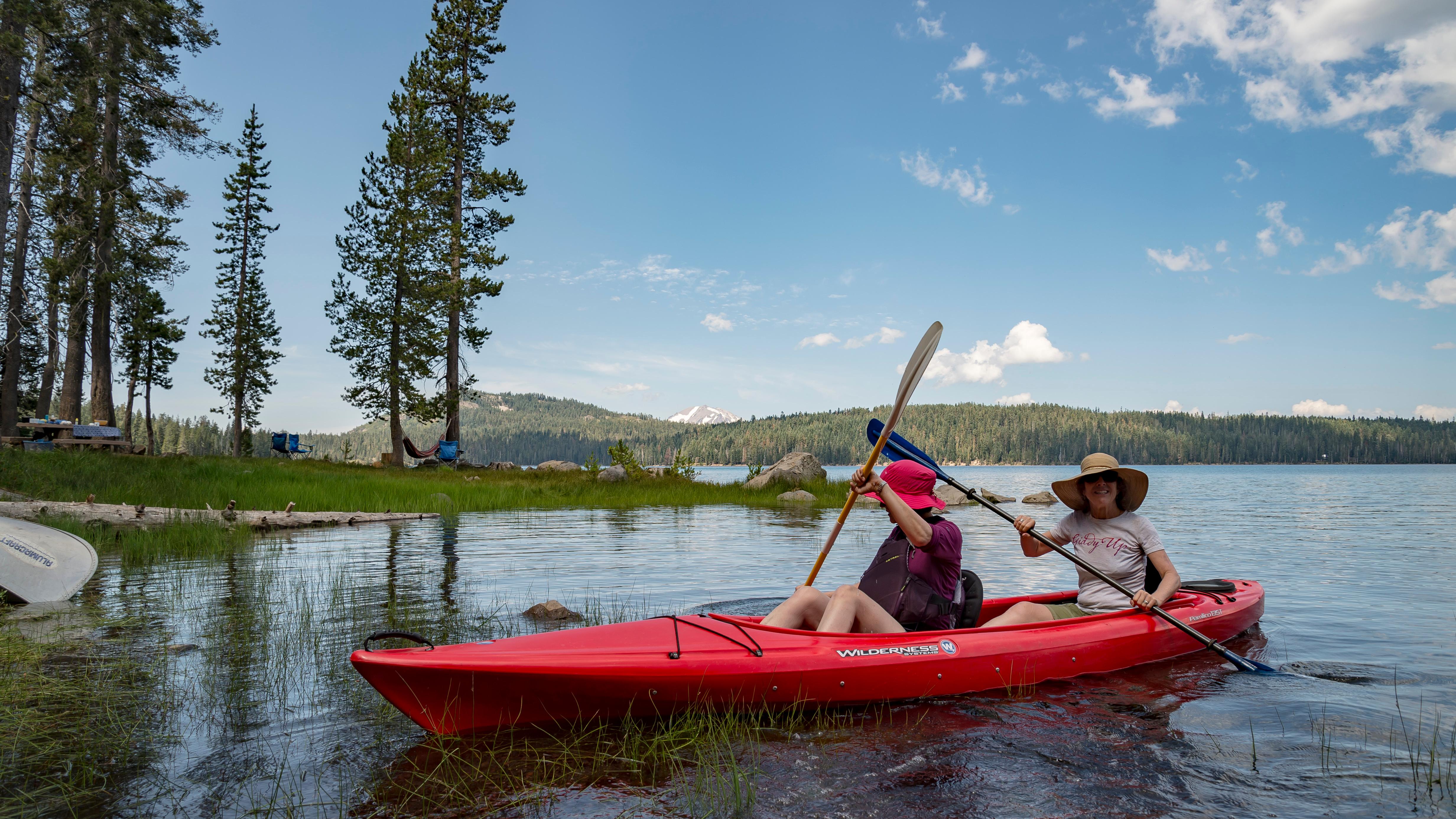 Two women sit in a red kayak on a lake backed by a snow-capped mountain