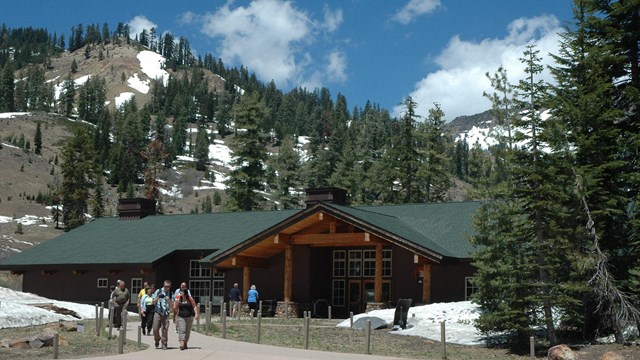 people approaching a large, cabin-like building with snowy hills behind