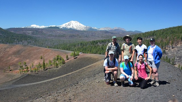 A group of hikers poses for a photo on the summit of a black ridge backed by a snow-capped volcano