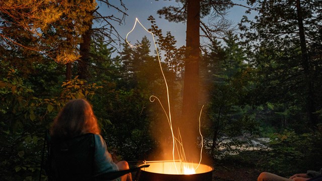 a fire in a metal fire ring at night, a woman sitting next to the fire, stars and trees overhead