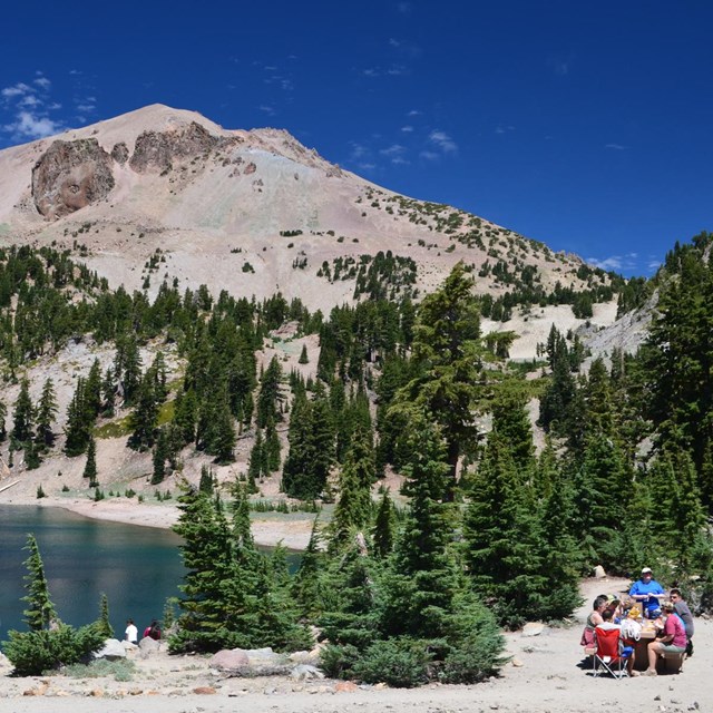 A group of people sitting at a picnic table on the edge of a mountain lake beneath a large volcano.