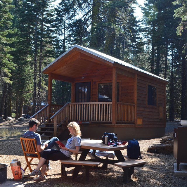 A man a woman sit and read outside of a small, wooden cabin